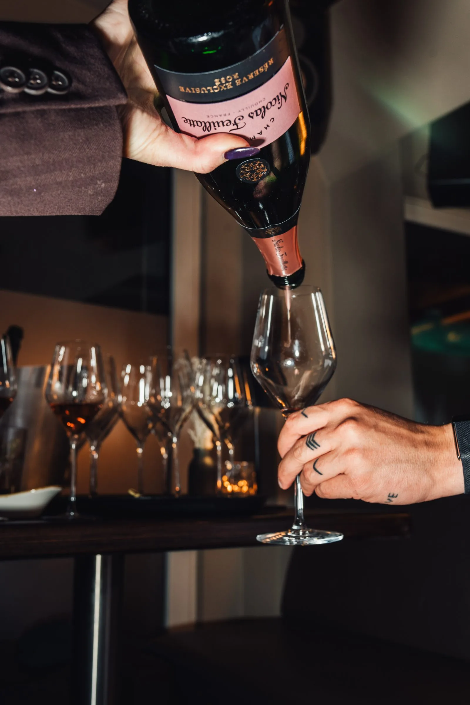 Rosé-Champagner wird an der Bar in ein Weinglas eingeschenkt, dahinter weitere Gläser im warmen Licht.